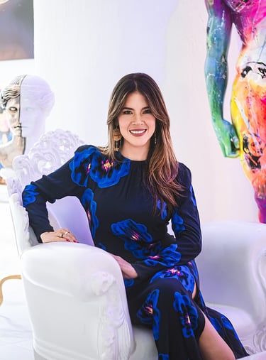 Natalia Devia sentada en un sillón blanco, rodeada de esculturas y arte colorido